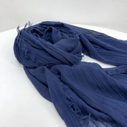 A navy blue lightweight linen scarf with a crinkled texture and frayed fringe edges, neatly swirled and laid out on a clean white surface.