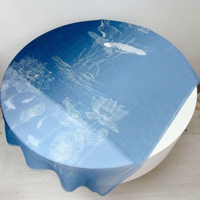 A lightweight, sheer scarf with a blue gradient background featuring delicate white fine-line illustrations of lotus flowers, pads, and swimming koi fish spread across a round table.