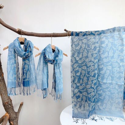 Three lightweight blue scarves with white floral leaf patterns displayed on wooden hangers and draped over a branch, featuring frayed fringe trim along the edges against a white backdrop.