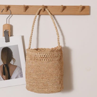 A natural beige handcrafted woven straw bucket bag with a braided shoulder strap and scalloped crocheted top trim, hanging from a wooden wall peg alongside a framed print against a plain white wall.