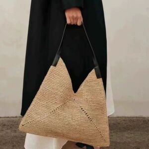 A person in a black coat holding a large, beige woven straw origami-style tote bag with a black leather shoulder strap.