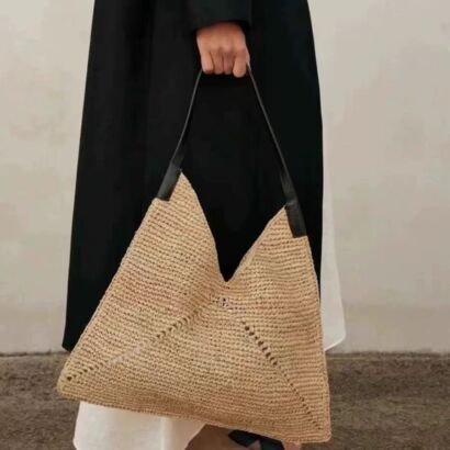 A person in a black coat holding a large, beige woven straw origami-style tote bag with a black leather shoulder strap.