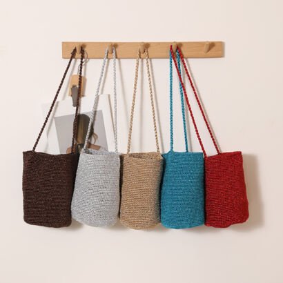 Five woven raffia mini crossbody bucket bags in dark brown, silver grey, beige, teal blue, and vibrant red hanging in a row from wooden wall pegs against a plain off-white wall.