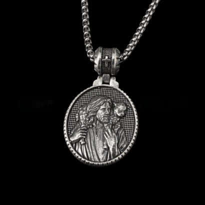 A detailed silver-tone metal pendant necklace hanging against a black background, featuring a round medallion with a high-relief carving of the Good Shepherd holding a lamb against a grid background, suspended from a matching chain via an ornate cross-accented bail.