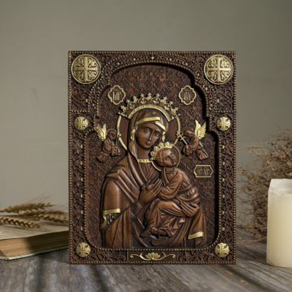 An upright rectangular wooden wall plaque featuring a detailed high-relief carving of the Madonna and Child in dark wood with gold-accented highlights and ornate patterned borders, displayed on a wooden surface with a book and a candle.