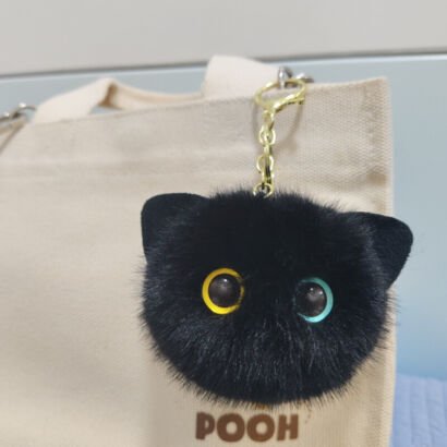 A round, fluffy black fur cat head keychain featuring cute pointed ears and odd-colored heterochromia eyes (one yellow, one blue), clipped to a cream canvas tote bag with a gold clasp.