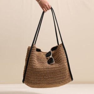 A handwoven rich brown straw hobo shoulder bag held by a hand, featuring dark black faux leather shoulder straps and side trim, with white-framed sunglasses tucked into the top opening against a neutral backdrop.