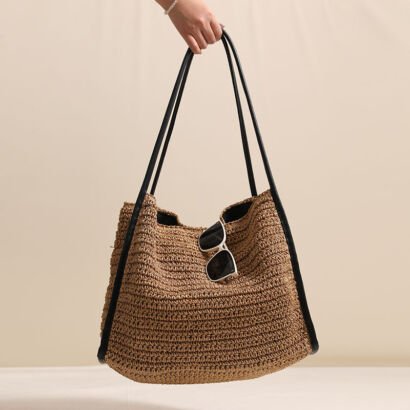 A handwoven rich brown straw hobo shoulder bag held by a hand, featuring dark black faux leather shoulder straps and side trim, with white-framed sunglasses tucked into the top opening against a neutral backdrop.