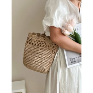 A woman in a white dress holding light pink tulips and a magazine while wearing a handwoven beige straw bucket shoulder bag featuring an openwork lattice band and a scalloped crocheted top border.