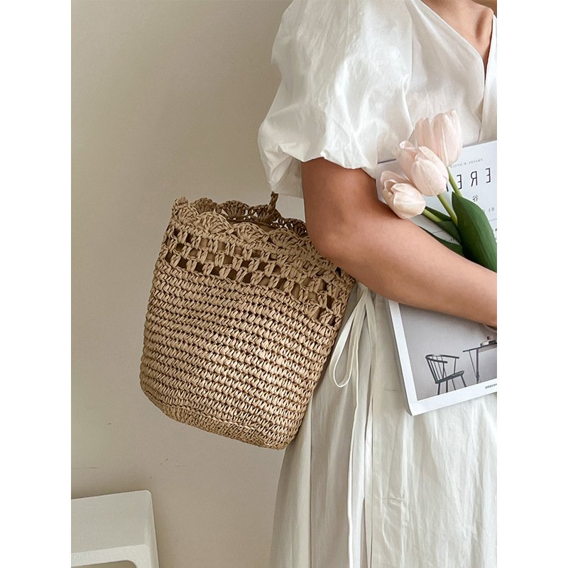 A woman in a white dress holding light pink tulips and a magazine while wearing a handwoven beige straw bucket shoulder bag featuring an openwork lattice band and a scalloped crocheted top border.