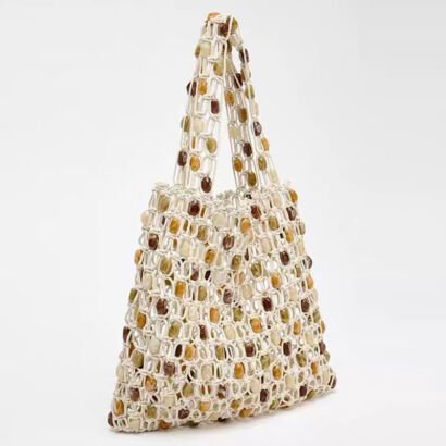 A handmade off-white macrame open-mesh net tote bag embellished with smooth, polished earth-tone stone beads in shades of brown, amber, and olive green, featuring matching woven handles against a plain white background.