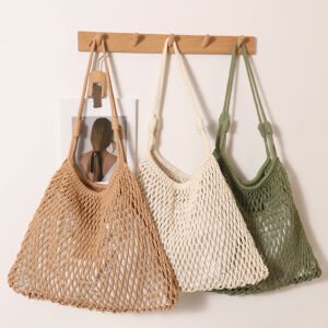 Three handmade open-mesh crochet net tote bags in beige, off-white, and sage green hanging side-by-side on a wooden wall peg against a clean white wall.