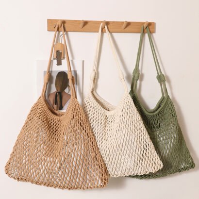 Three handmade open-mesh crochet net tote bags in beige, off-white, and sage green hanging side-by-side on a wooden wall peg against a clean white wall.
