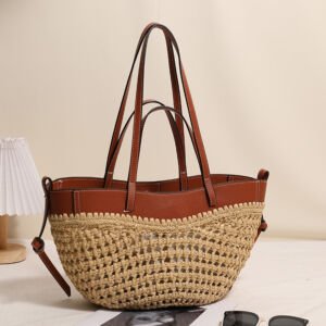 A handwoven natural straw basket tote bag with a rich brown faux leather upper rim, double sets of leather shoulder straps, and side tie details sitting on a table next to sunglasses and a lamp.