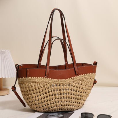 A handwoven natural straw basket tote bag with a rich brown faux leather upper rim, double sets of leather shoulder straps, and side tie details sitting on a table next to sunglasses and a lamp.