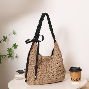 A natural beige handwoven straw hobo shoulder bag featuring a black scrunched fabric shoulder strap tied in a delicate ribbon bow at the side, displayed on a round white table next to a coffee cup and sunglasses.