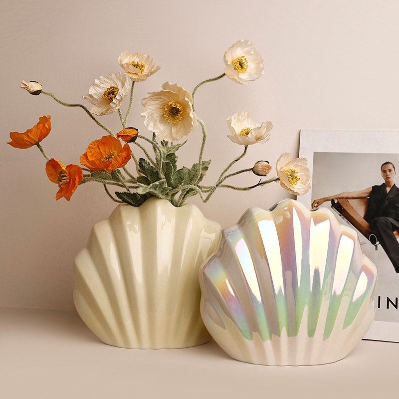 Two clam seashell-shaped ceramic vases, one soft cream and one with an iridescent mother-of-pearl sheen, holding white and orange poppies