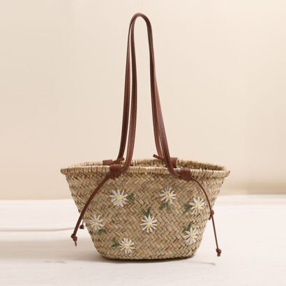 A handwoven natural straw basket tote bag embroidered with white floral daisy motifs, featuring long brown leather shoulder straps and side drawstring ties against a soft neutral background.