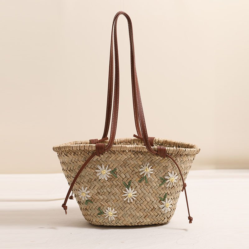 A handwoven natural straw basket tote bag embroidered with white floral daisy motifs, featuring long brown leather shoulder straps and side drawstring ties against a soft neutral background.