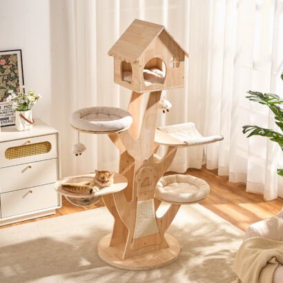 A tall, tree-shaped natural wooden cat tower featuring a house-shaped top condo with a cushion, two plush round perches, a fabric hammock, a clear acrylic bowl hammock with an orange tabby cat lying inside, and hanging plush toys, set in a bright modern living room.