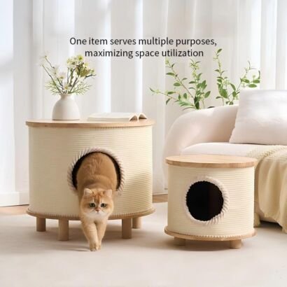 Two cylindrical beige cream cat condo hideaways with light wood tabletops and wooden legs sitting on a floor next to a white sofa, with an orange tabby cat stepping out of the larger condo's circular entryway.