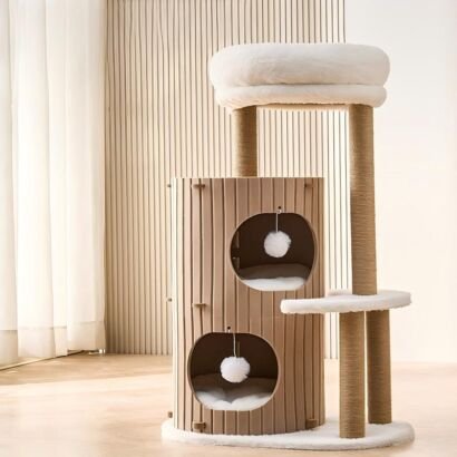 A modern neutral-toned cat tree featuring a warm beige slatted two-story cylindrical condo with soft white cushions and dangling pom-poms, sisal-wrapped scratching posts, a small side step, and a thick plush top round bed set against a bright interior wall.
