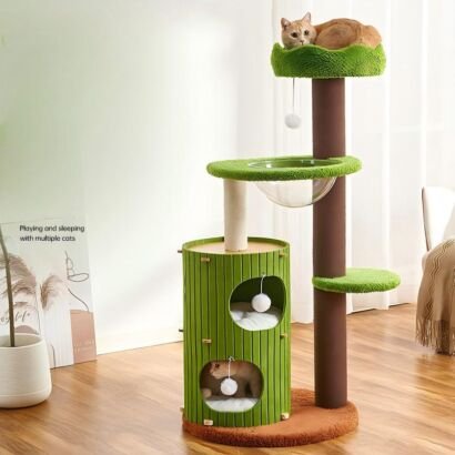 A tall, nature-inspired multi-level cat tree featuring vibrant green plush perches, a tall brown main post, a green cylindrical two-story cat condo, a transparent acrylic bowl hammock, dangling plush pom-poms, and an orange tabby cat resting at the top perch.