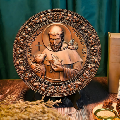 A round bronze-finish decorative wall plaque standing upright on a stand against a dark green curtain and wooden surface, featuring a high-relief carving of Saint Francis holding a lamb and a bird, encircled by text reading "SAINT FRANCIS PROTECT US" and an ornate grape vine border.
