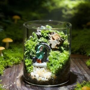 A cylindrical clear glass terrarium filled with vibrant green moss, natural rockwork, a gravel pathway, and a tiny figurine in a green raincoat standing outside a arched door set against a lush forest backdrop.