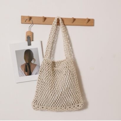 A beige handwoven open-mesh crochet net tote bag hanging on a wooden wall peg, featuring integrated open-weave shoulder straps against a plain neutral wall.