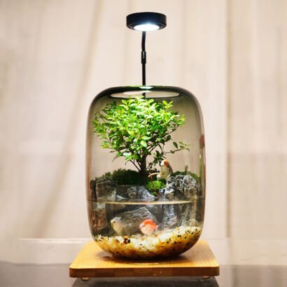 A rounded glass capsule paludarium featuring an underwater aquascape with small goldfish swimming over gravel, topped by a lush miniature tree, mossy rockwork, and a fishing cat figurine, illuminated by an overhead LED light.