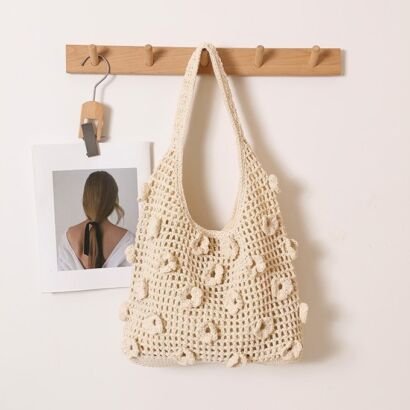 A cream-white handwoven crochet shoulder tote bag with 3D popcorn-stitch floral embellishments scattered across an open-work mesh knit, hanging on a wooden wall rack.