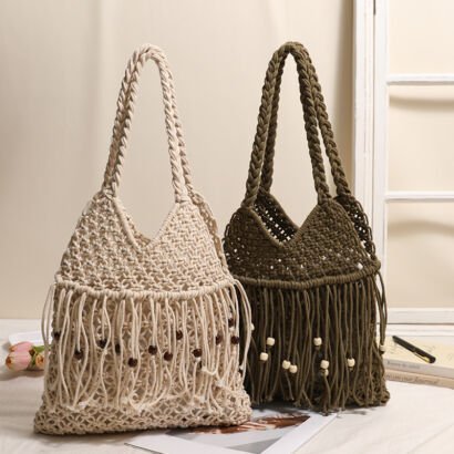 Two handmade macrame straw shoulder bags in cream and olive green sitting side-by-side on a light surface, featuring thick braided rope handles, long fringe trim, and decorative wooden bead accents.