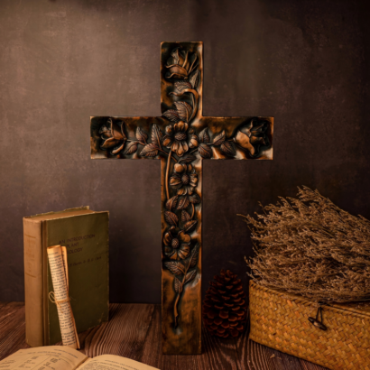 A dark wood carved wall cross standing upright on a rustic wooden table next to vintage books, a scroll, a pinecone, and a woven basket of dried branches, featuring an intricate high-relief carving of blossoming roses and floral vines trailing across the cross.