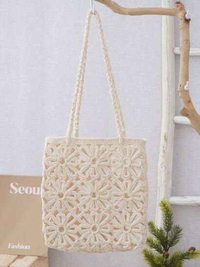 A cream-white handwoven crochet tote bag featuring an intricate open-work floral daisy pattern, hanging from long braided shoulder straps against a neutral wall backdrop.