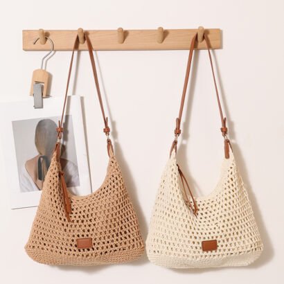 Two handwoven crochet straw hobo shoulder bags in natural khaki and cream white hanging side-by-side on a wooden wall peg, each featuring brown leather shoulder straps and a small faux leather logo patch on the front.