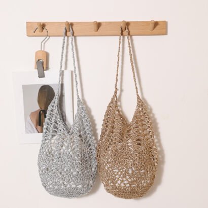 Two glittery crochet open-work straw hobo bags—one in metallic silver and one in metallic gold—hanging side-by-side on a wooden wall peg, featuring long matching woven shoulder straps.