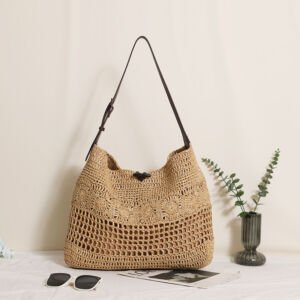 A natural beige handwoven straw hobo shoulder bag with a dark brown leather strap and toggle closure, featuring a hollow-out mesh pattern, displayed next to sunglasses and a vase on a light tabletop.