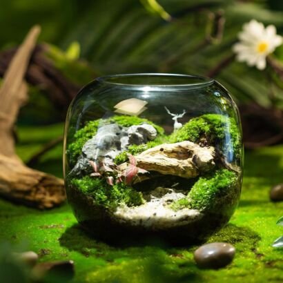 A rounded clear glass terrarium bowl set on a vibrant green mossy surface, featuring vibrant moss hills, natural rocks, driftwood, red foliage, and a tiny white deer figurine.