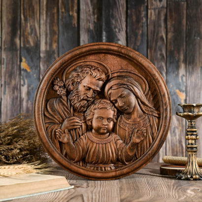 A round carved wooden wall plaque standing upright on a rustic wooden table against a dark wood background, featuring a high-relief sculpture of the Holy Family with Saint Joseph, the Virgin Mary, and the Child Jesus.