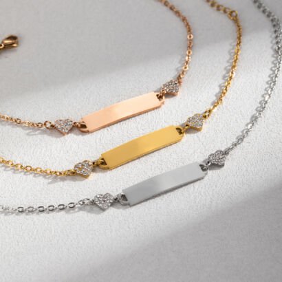 Three delicate chain bracelets in rose gold, yellow gold, and silver finishes, each featuring a blank rectangular bar plate flanked by two pavé crystal heart connectors on a textured light surface.