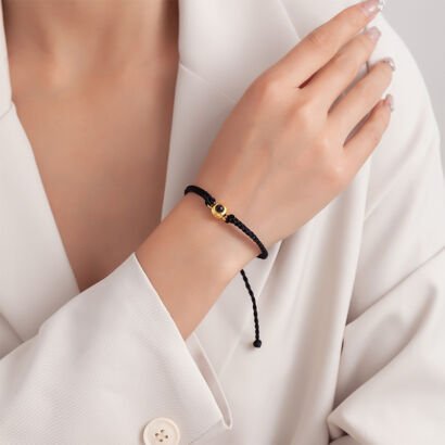 A hand resting on a white blazer sleeve wearing a delicate black braided string bracelet featuring a central round gold bead with a dark projection lens.