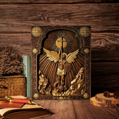 A rectangular carved wooden wall plaque standing upright on a rustic wooden table against a dark wood background, featuring a high-relief sculpture of Archangel Michael with golden wings holding a cruciform staff beneath a radiant sunburst arch, surrounded by ornate patterned borders and corner crosses.