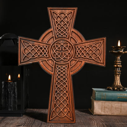 Ornately carved dark wood Celtic cross with intricate knotwork patterns and central ring setting on a tabletop next to a glowing candle and vintage books