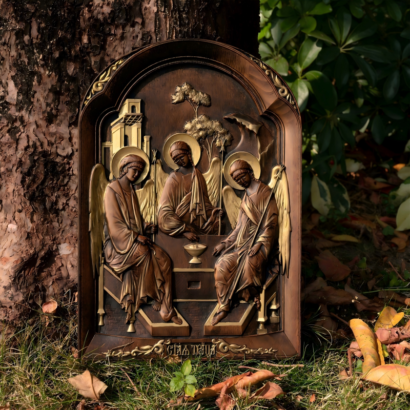 An arched carved wooden wall plaque standing outdoors against a tree trunk and grass, featuring a high-relief sculptural depiction of Andrei Rublev's Holy Trinity with three winged angels seated around a table.