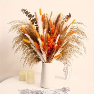 A full, textured dried floral arrangement featuring neutral pampas grass, white plumes, and vibrant yellow and orange bunny tail accents displayed in a white ceramic vase alongside small candles on a white tabletop.