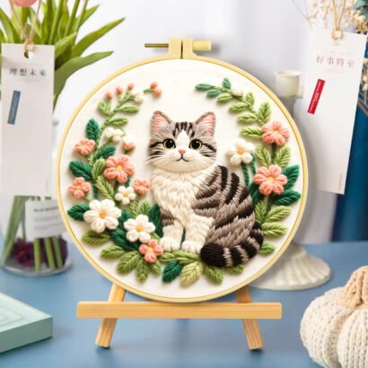 A round wooden embroidery hoop set on a small easel, displaying a 3D textured embroidered grey striped tabby cat sitting inside a circular wreath of green leaves, pink flowers, and white blossoms against a light fabric background.