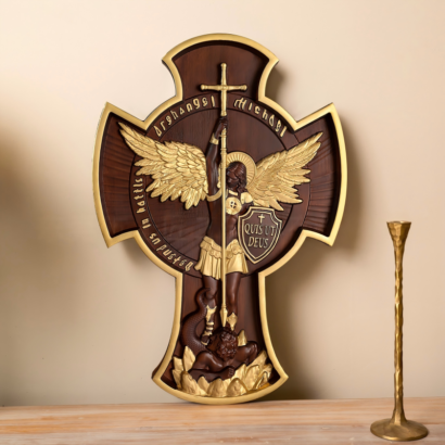 An ornate wooden cross wall plaque standing upright on a wooden table next to a brass candlestick, featuring a dark wood and gold-accented carved relief of Archangel Michael with wings and a sword subduing a figure below, inscribed with "QUIS UT DEUS".
