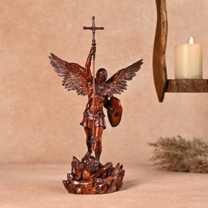 A detailed dark bronze-finish figurine of Archangel Michael standing triumphantly with outstretched wings, holding a sword-cross aloft and a shield while subduing Satan on a rocky base, set against a neutral background with a lit candle.
