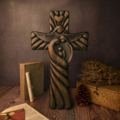 A dark wood carved wall cross standing upright on a rustic wooden table surrounded by vintage books, a rolled scroll, a pinecone, and a woven box, featuring a stylized sculptural relief of the Holy Family with intertwined figures.